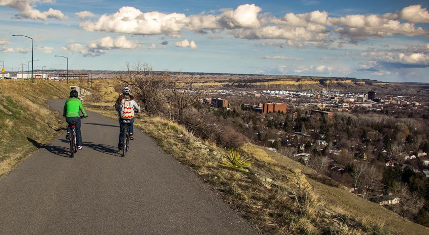 Two bicyclists riding away from the camera downhill on a bike path with a city in the distance