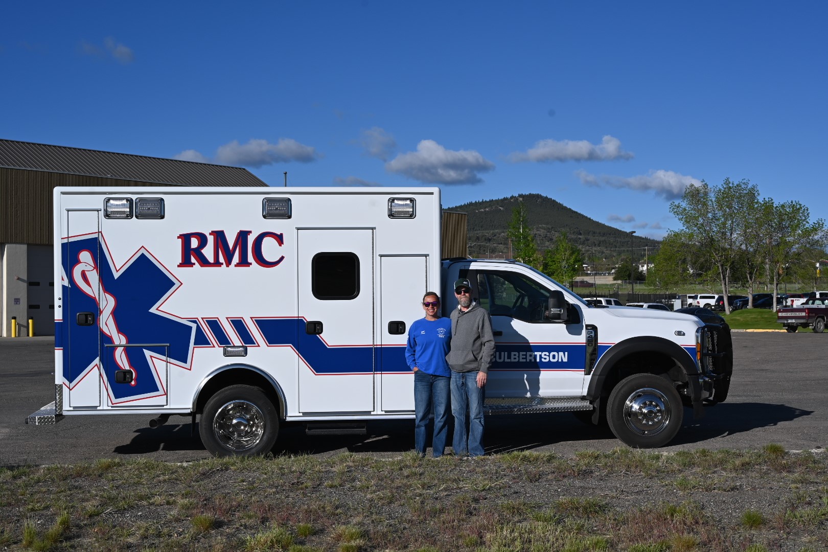 Two people standing in front of a white RMC ambulance parked outdoors near a building and trees under a clear blue sky.